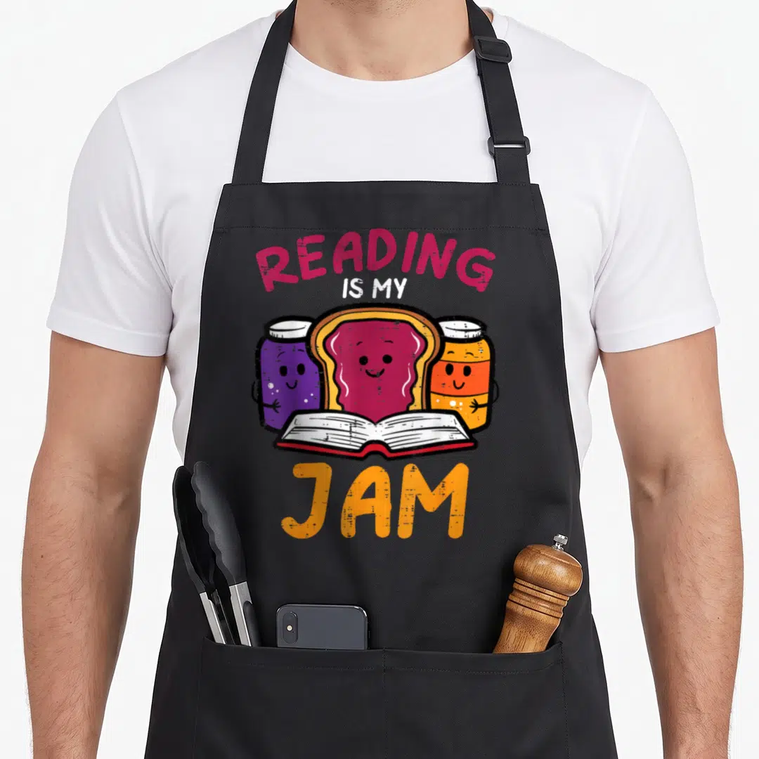 Jam reading books apron - Cartoon toast between purple and orange jam jars behind open book with READING IS MY JAM text — shown on a black cotton canvas apron against a white background