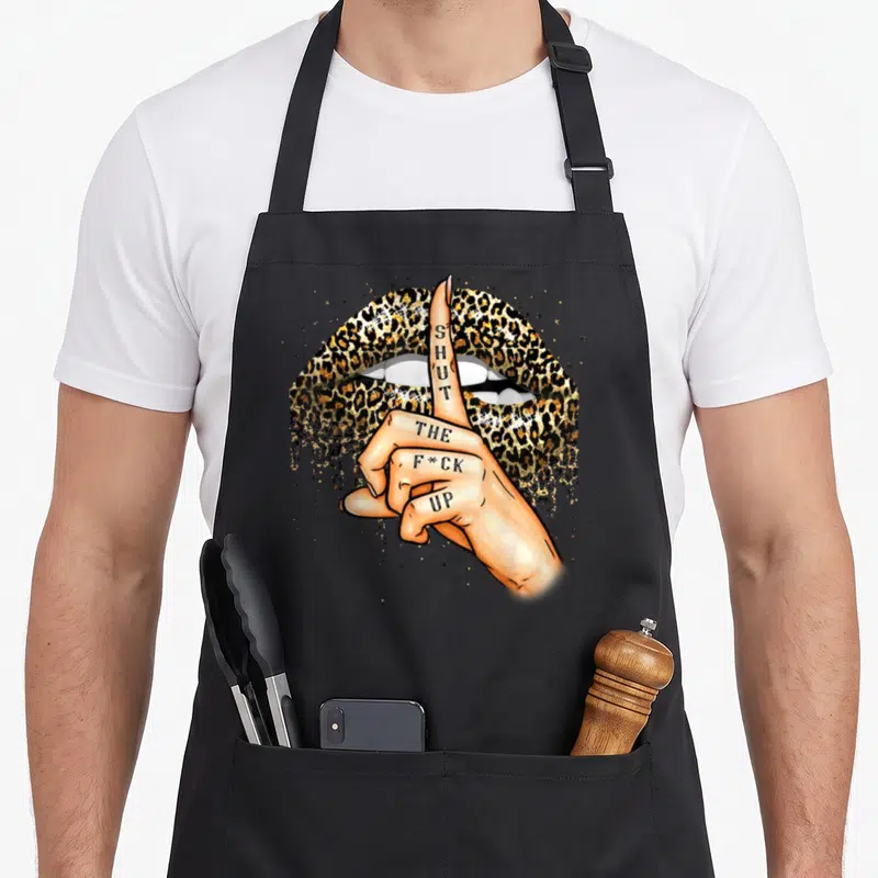 Shut leopard apron - Hand with tattoo text SHUT THE F*CK UP over gold leopard lips — shown on a black cotton canvas apron against a white background