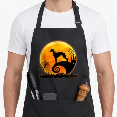 Scary dog apron - Black Whippet silhouette on swirling hill against orange moon with bats and castle — shown on a black cotton canvas apron against a white background