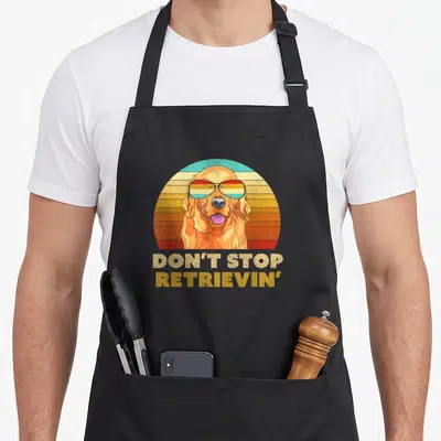 Stop golden retriever apron - Golden retriever in striped sunglasses under orange and teal sunburst with stacked text — shown on a black cotton canvas apron against a white background