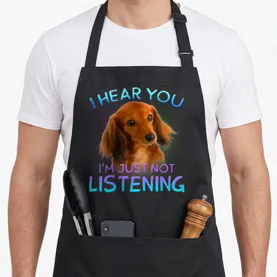 Haired dachshund apron - Brown dachshund head photo with I HEAR YOU I'M JUST NOT LISTENING text in blue and purple — shown on a black cotton canvas apron against a white background