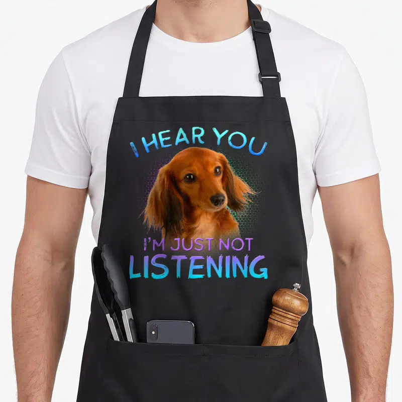 Haired dachshund apron - Brown dachshund head photo with I HEAR YOU I'M JUST NOT LISTENING text in blue and purple — shown on a black cotton canvas apron against a white background