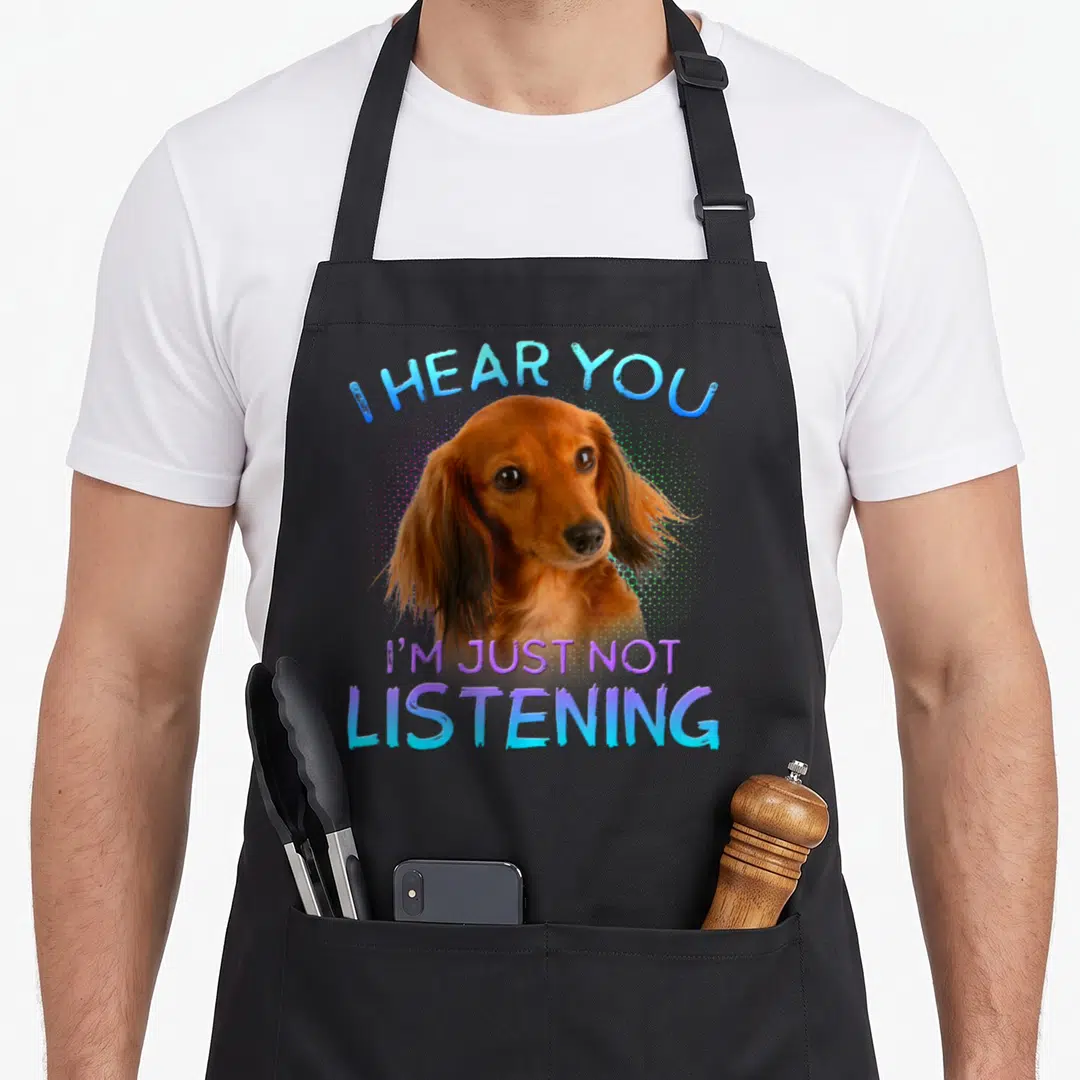 Haired dachshund apron - Brown dachshund head photo with I HEAR YOU I'M JUST NOT LISTENING text in blue and purple — shown on a black cotton canvas apron against a white background