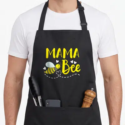 Mama bee apron - Yellow MAMA Bee text with cartoon bee, white hearts, and dashed flight line — shown on a black cotton canvas apron against a white background