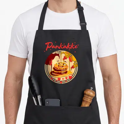 Cake pancake apron - Pankakke text above cartoon pancakes with syrup bottles and red hearts in yellow red brown — shown on a black cotton canvas apron against a white background