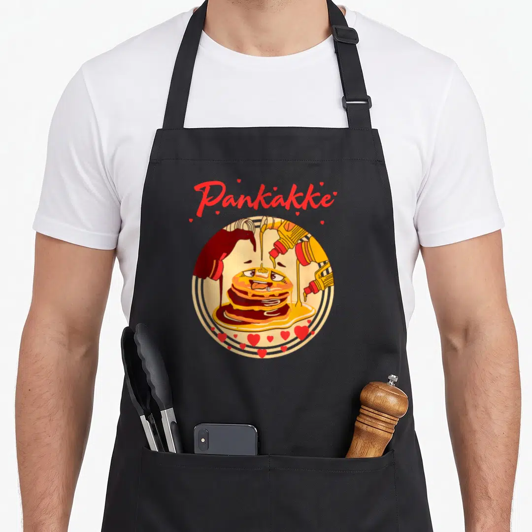 Cake pancake apron - Pankakke text above cartoon pancakes with syrup bottles and red hearts in yellow red brown — shown on a black cotton canvas apron against a white background