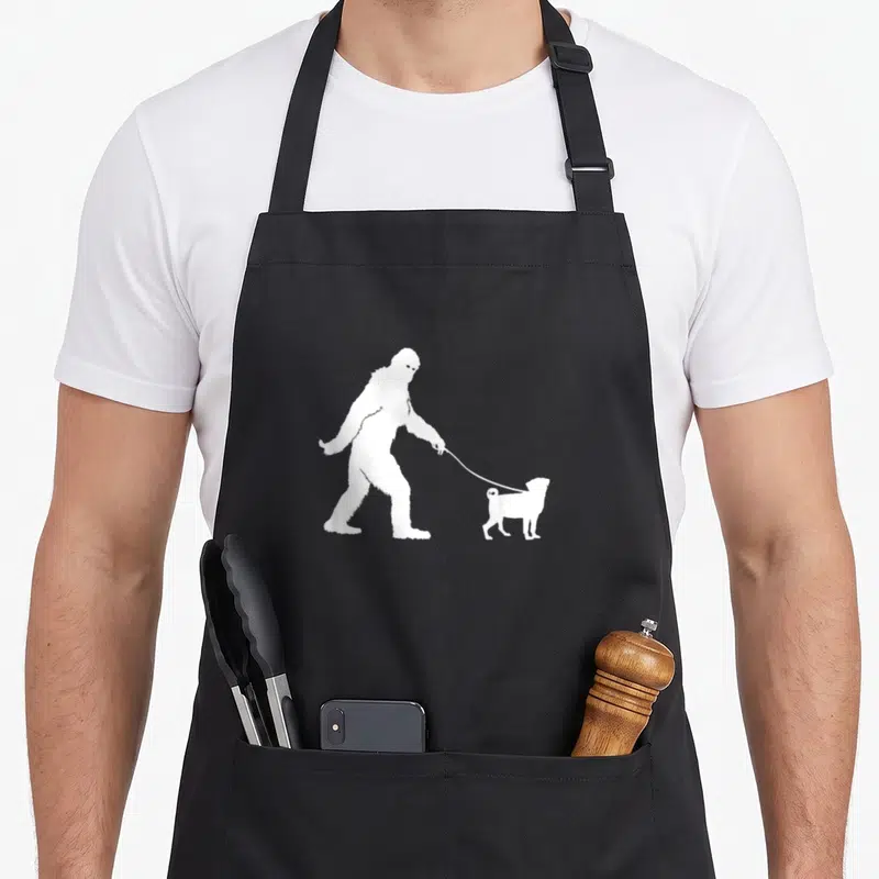 Sasquatch walking apron - White bigfoot silhouette walking a small pug dog on a leash — shown on a black cotton canvas apron against a white background