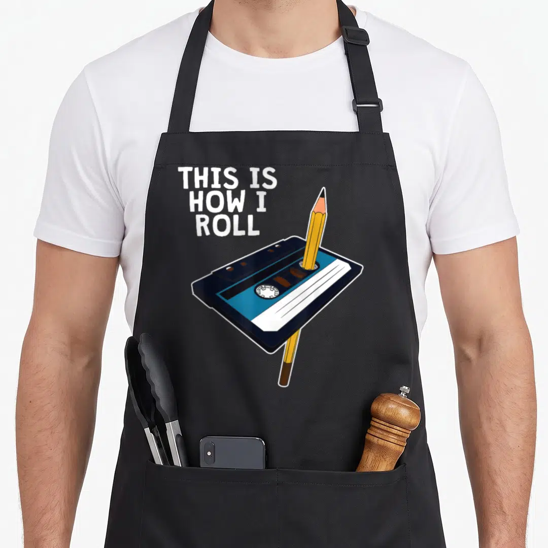 Tape 70s apron - Cassette tape with pencil and stacked text THIS IS HOW I ROLL in white and blue — shown on a black cotton canvas apron against a white background