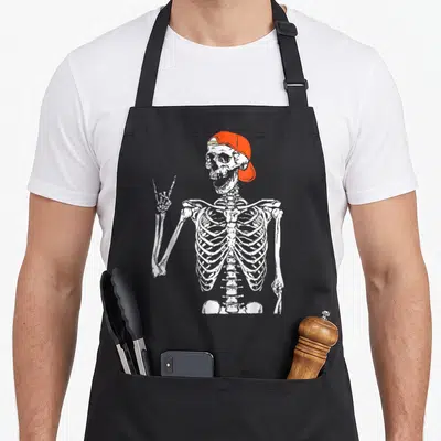 Rocker skeleton apron - White skeleton wearing orange cap making rock-on hand gesture on black background — shown on a black cotton canvas apron against a white background
