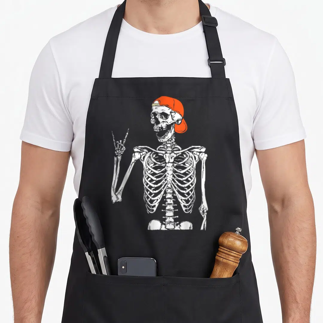 Rocker skeleton apron - White skeleton wearing orange cap making rock-on hand gesture on black background — shown on a black cotton canvas apron against a white background