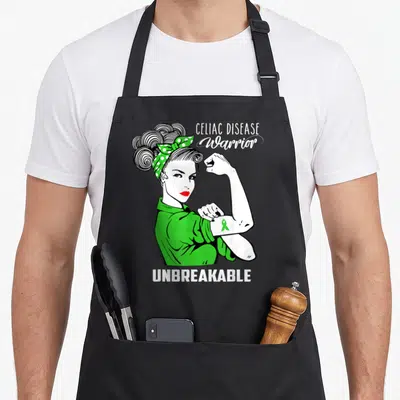 Disease celiac awareness apron - Cartoon woman flexing arm with green ribbon and Celiac Disease Warrior Unbreakable text — shown on a black cotton canvas apron against a white background