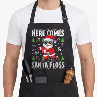 Santa come and get it apron - Cartoon Santa flossing with HERE COMES SANTA FLOSS text and candy cane pattern — shown on a black cotton canvas apron against a white background