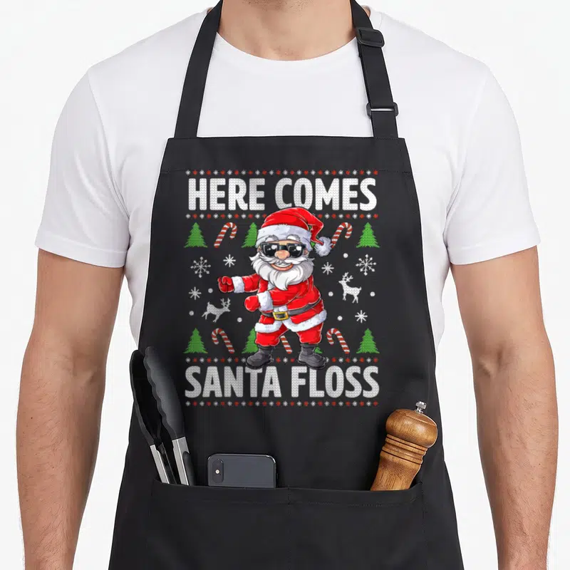 Santa come and get it apron - Cartoon Santa flossing with HERE COMES SANTA FLOSS text and candy cane pattern — shown on a black cotton canvas apron against a white background