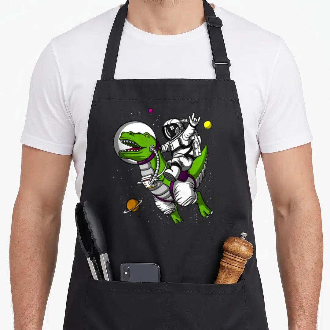 Astronaut science apron - Cartoon astronaut riding green dinosaur with three colorful planets in space — shown on a black cotton canvas apron against a white background