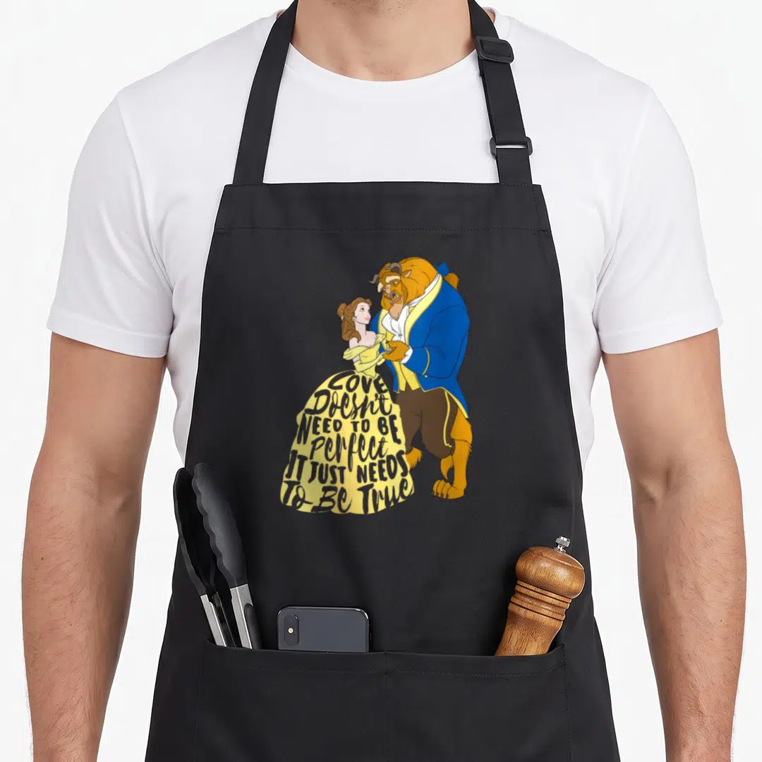 Funny key lover apron - Beast and Belle standing together with text on yellow dress reading LOVE Doesn't NEED TO BE — shown on a black cotton canvas apron against a white background