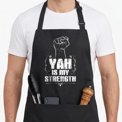 Strength hebrew aprons - White distressed clenched fist above stacked YAH IS MY STRENGTH text with swirls — shown on a black cotton canvas apron against a white background