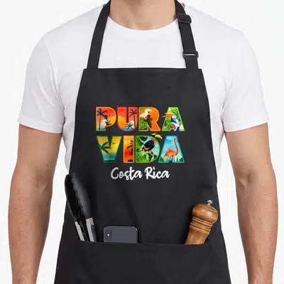Pura travel aprons - Pura Vida letters filled with toucan, flamingo, and palm tree scenes with Costa Rica script below — shown on a black cotton canvas apron against a white background