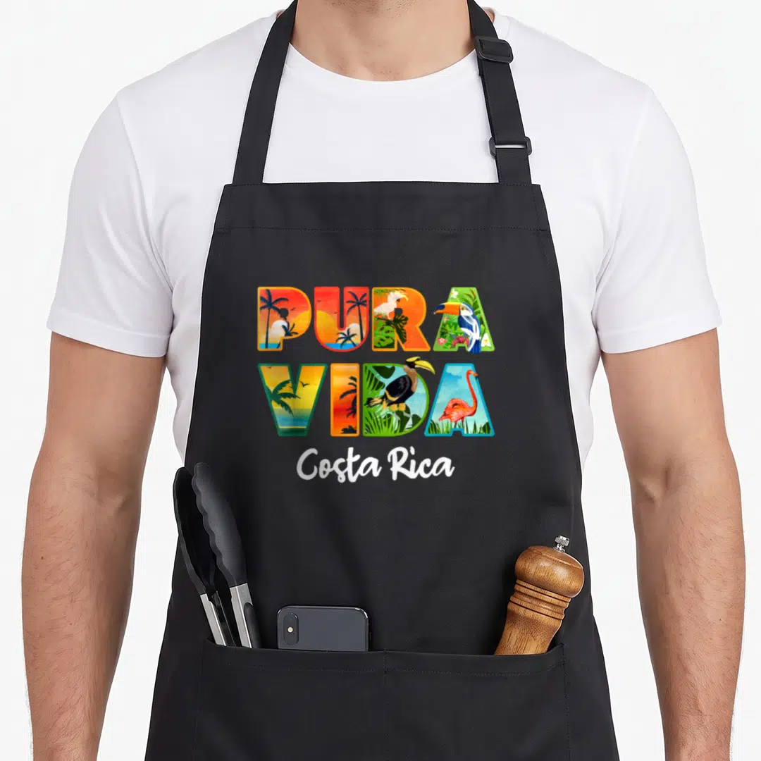 Pura travel aprons - Pura Vida letters filled with toucan, flamingo, and palm tree scenes with Costa Rica script below — shown on a black cotton canvas apron against a white background