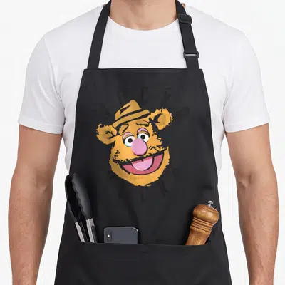 Wocka bear apron - Fozzie Bear head with arched WOCKA text in yellow and pink cartoon style — shown on a black cotton canvas apron against a white background