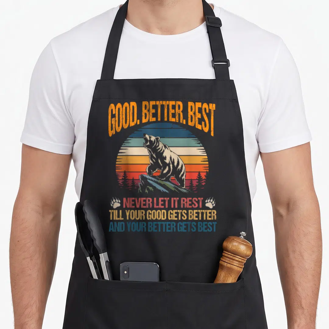 Better bear apron - Retro bear on striped sun with good better best never let it rest text and paw prints — shown on a black cotton canvas apron against a white background