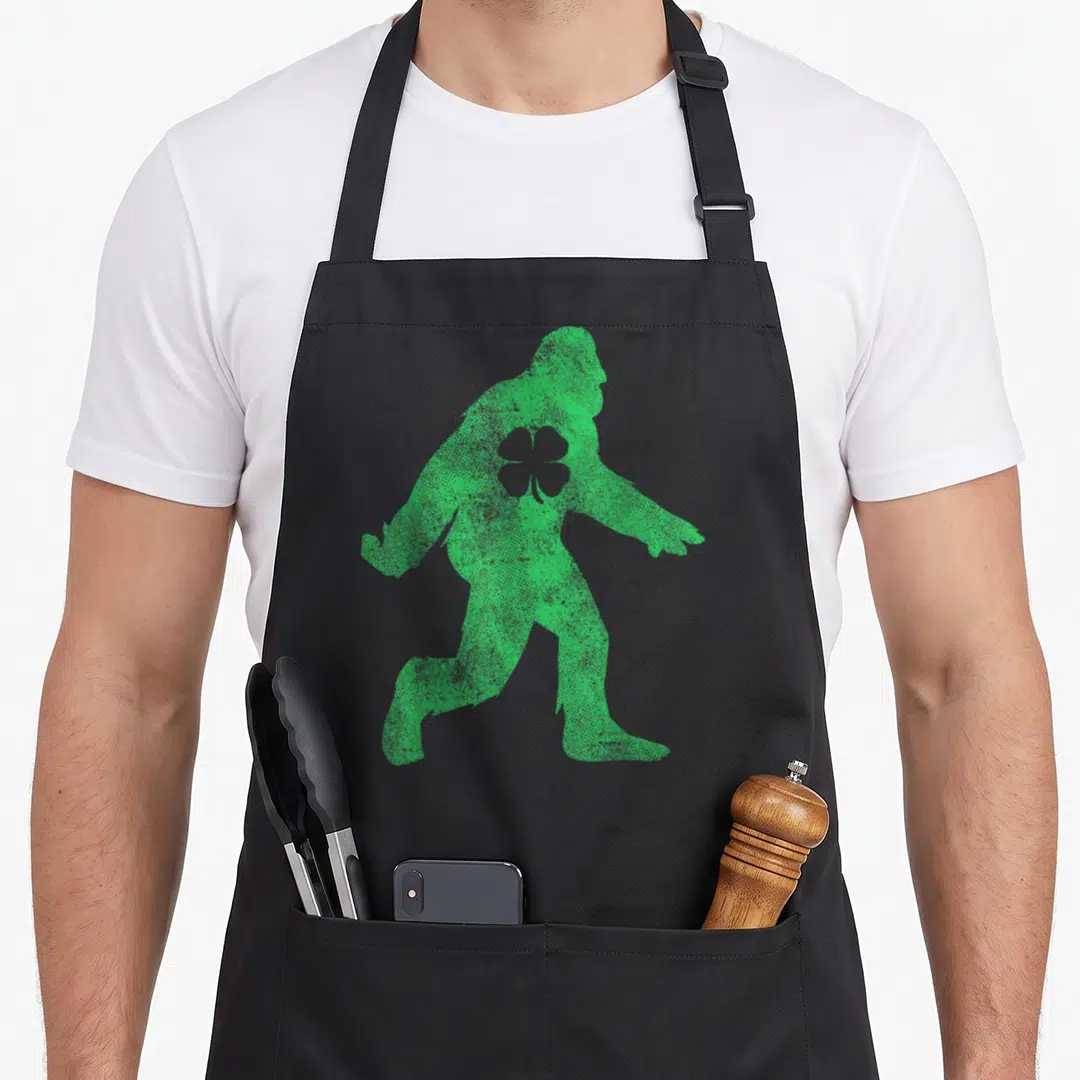 Sasquatch st patrick day aprons - Green distressed bigfoot silhouette with shamrock on chest — shown on a black cotton canvas apron against a white background