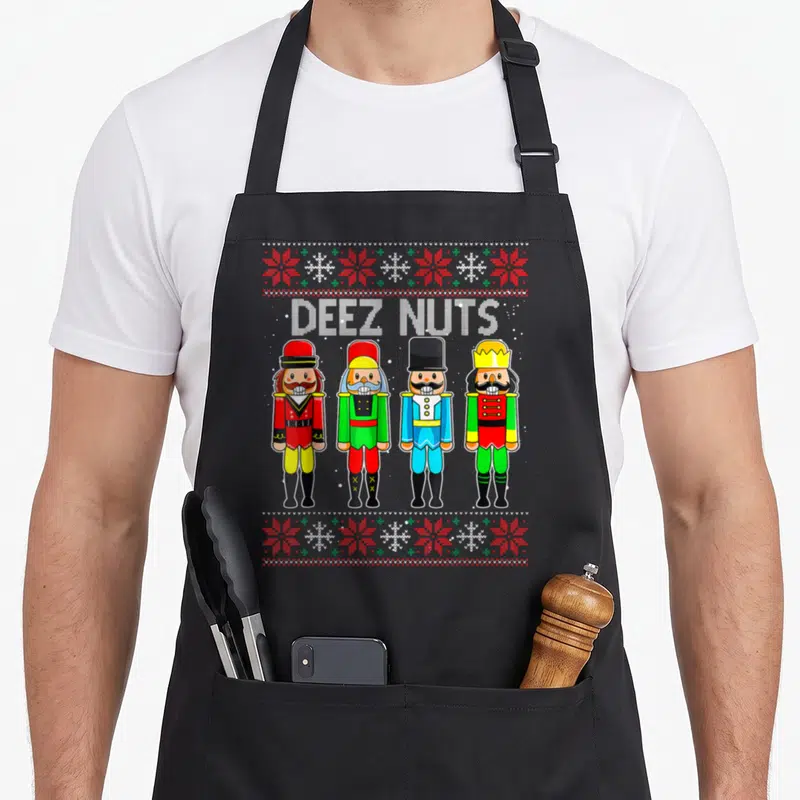 Deez xmas apron - DEEZ NUTS text above four cartoon nutcrackers flanked by red and green snowflake rows — shown on a black cotton canvas apron against a white background