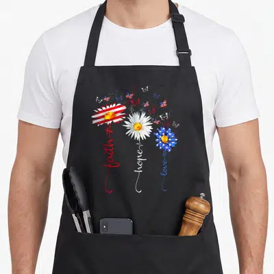 Faith 4th of july apron - Three daisies with red white and blue flag petals and stems reading faith hope love — shown on a black cotton canvas apron against a white background