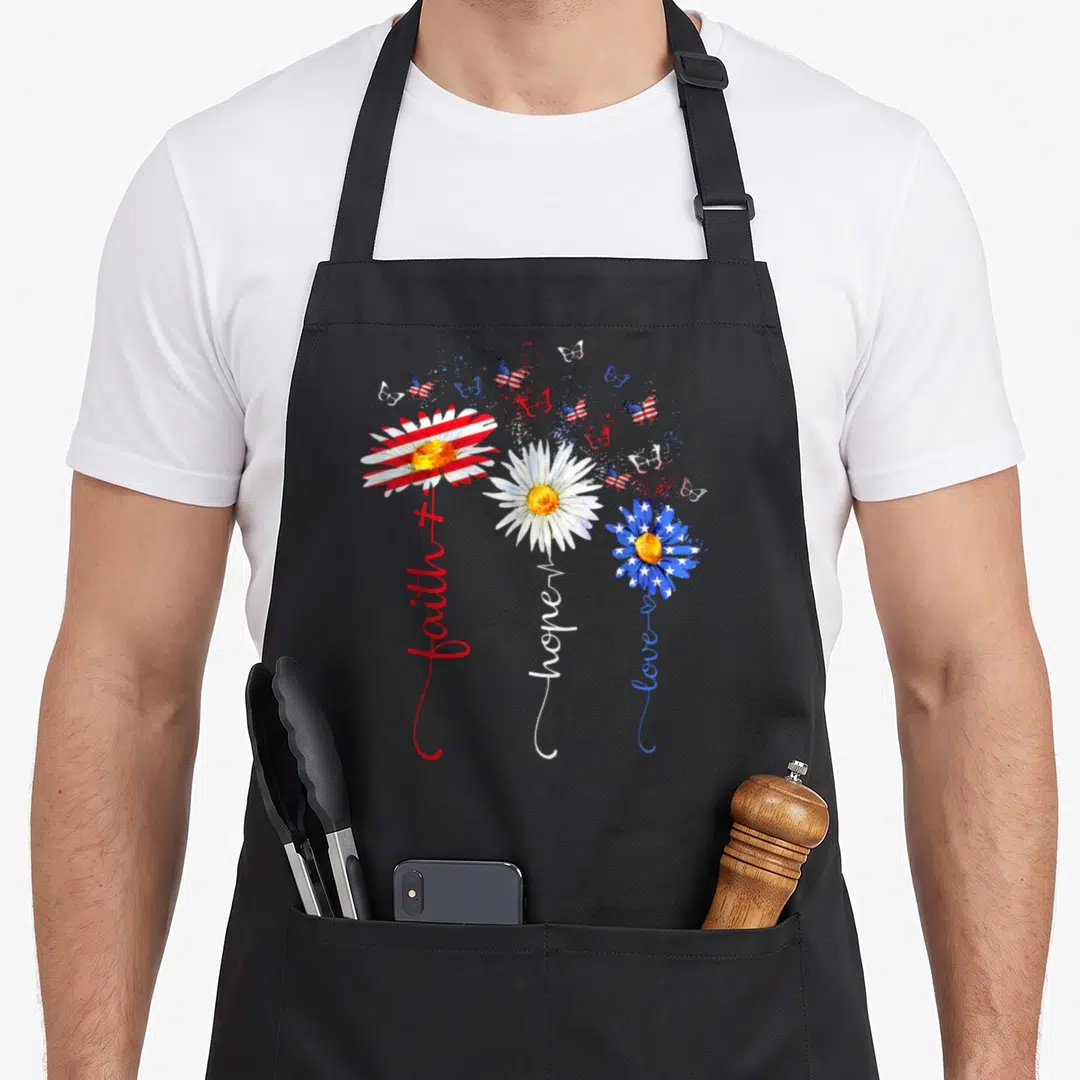 Faith 4th of july apron - Three daisies with red white and blue flag petals and stems reading faith hope love — shown on a black cotton canvas apron against a white background