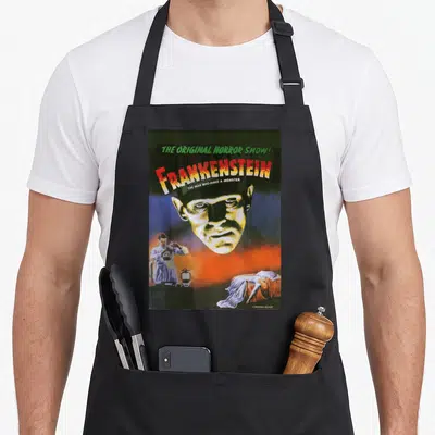 Monster distressed apron - Retro Frankenstein movie poster print with stacked text and character scenes in green, red — shown on a black cotton canvas apron against a white background
