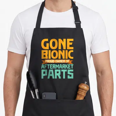 Bionic aftermarket parts of an apron - Orange and mint green stacked text reading Gone Bionic Proud Owner — shown on a black cotton canvas apron against a white background