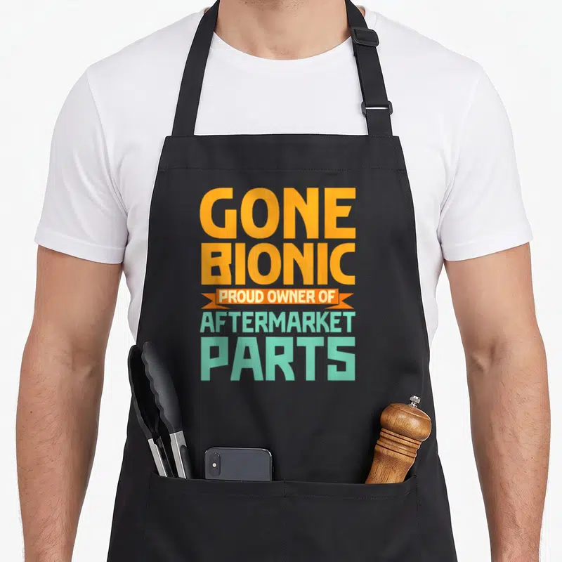 Bionic aftermarket parts of an apron - Orange and mint green stacked text reading Gone Bionic Proud Owner — shown on a black cotton canvas apron against a white background