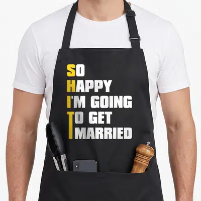 Sarcastic funny wedding aprons - Stacked yellow and white text spelling SHIT vertically within SO HAPPY I'M GOING — shown on a black cotton canvas apron against a white background