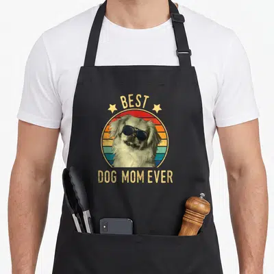 Pekingese mother's day aprons - Dog wearing sunglasses inside yellow red blue beige striped circle with BEST DOG MOM — shown on a black cotton canvas apron against a white background