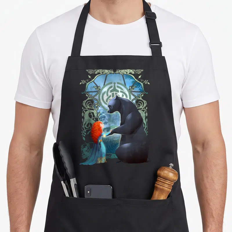 Brave bear apron - Black bear and red-haired girl in blue dress facing each other inside green ornamental frame — shown on a black cotton canvas apron against a white background