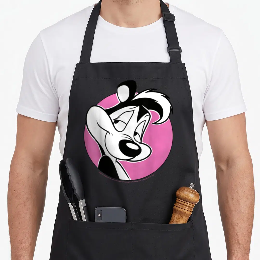 Pew le apron - Cartoon skunk head and shoulders centered on a pink circle background — shown on a black cotton canvas apron against a white background