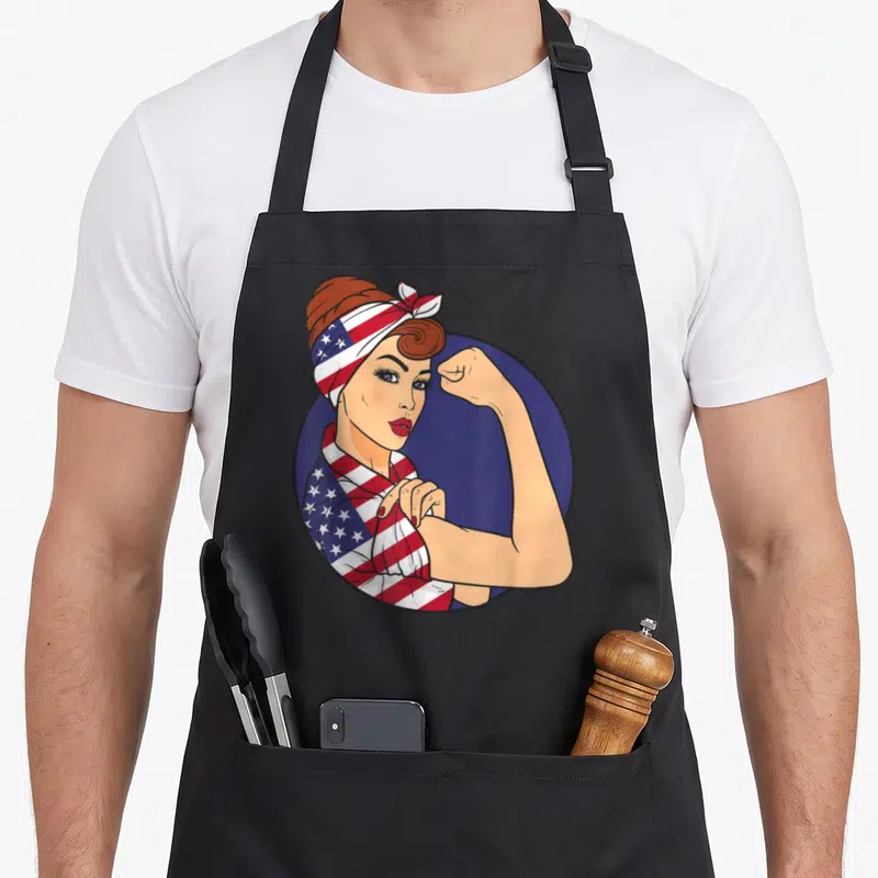 Riveter american flag apron - Cartoon woman flexing arm wearing US flag patterned shirt and headband inside blue circle — shown on a black cotton canvas apron against a white background