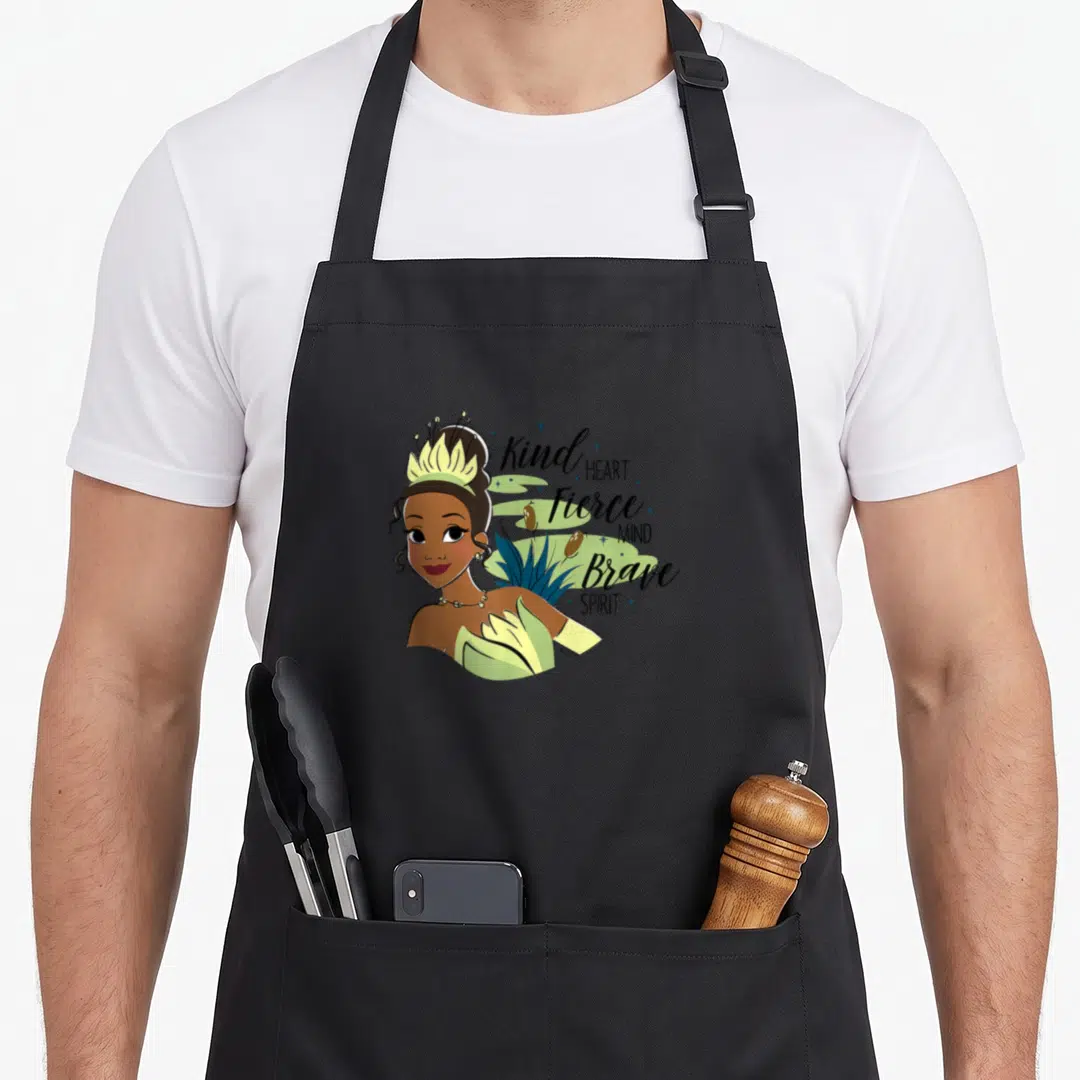 Kind princess apron - Cartoon woman with leaf crown beside Kind HEART Fierce MIND Brave SPIRIT text and reeds — shown on a black cotton canvas apron against a white background