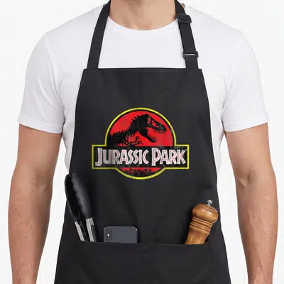 Original distressed apron - Tyrannosaurus Rex silhouette in red circle withJurassic Park text on yellow rectangle — shown on a black cotton canvas apron against a white background