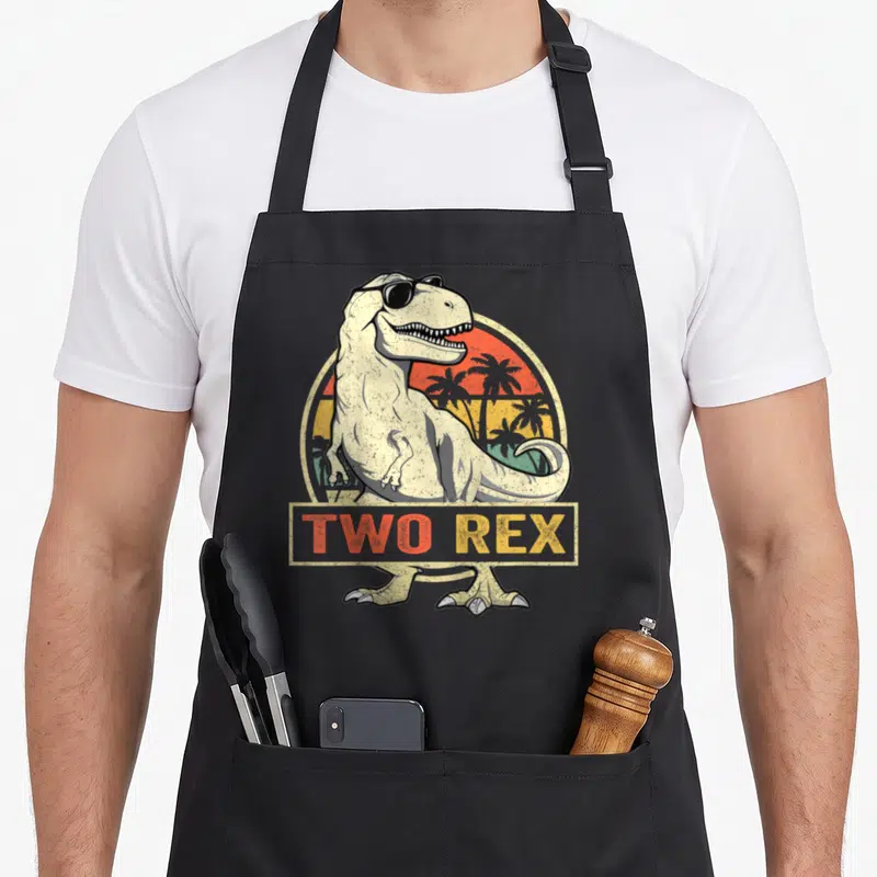 2nd t rex apron - Retro T-Rex with sunglasses and TWO REX text over a sunset — shown on a black cotton canvas apron against a white background