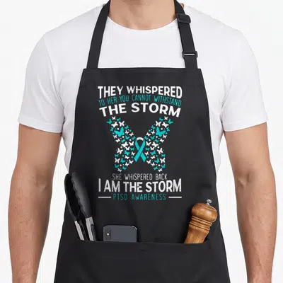 Posttraumatic butterfly apron - Distressed text I AM THE STORM with teal ribbon and butterfly cluster — shown on a black cotton canvas apron against a white background