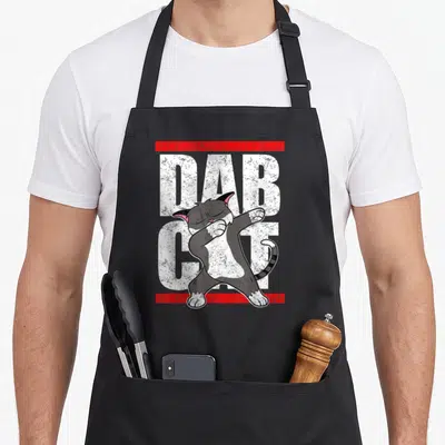Dab cat apron - Grey cartoon cat dabbing over white DAB CAT text with red bars — shown on a black cotton canvas apron against a white background