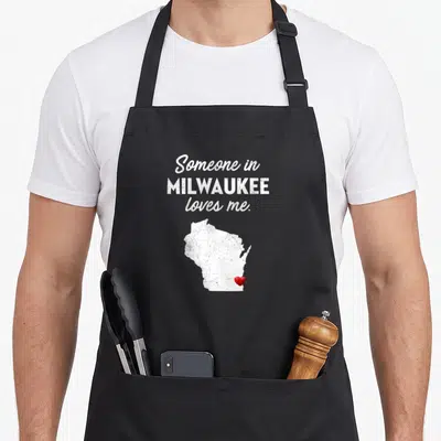 Someone milwaukee apron - Distressed white Wisconsin map with red heart and text reading Someone in MILWAUKEE loves me — shown on a black cotton canvas apron against a white background