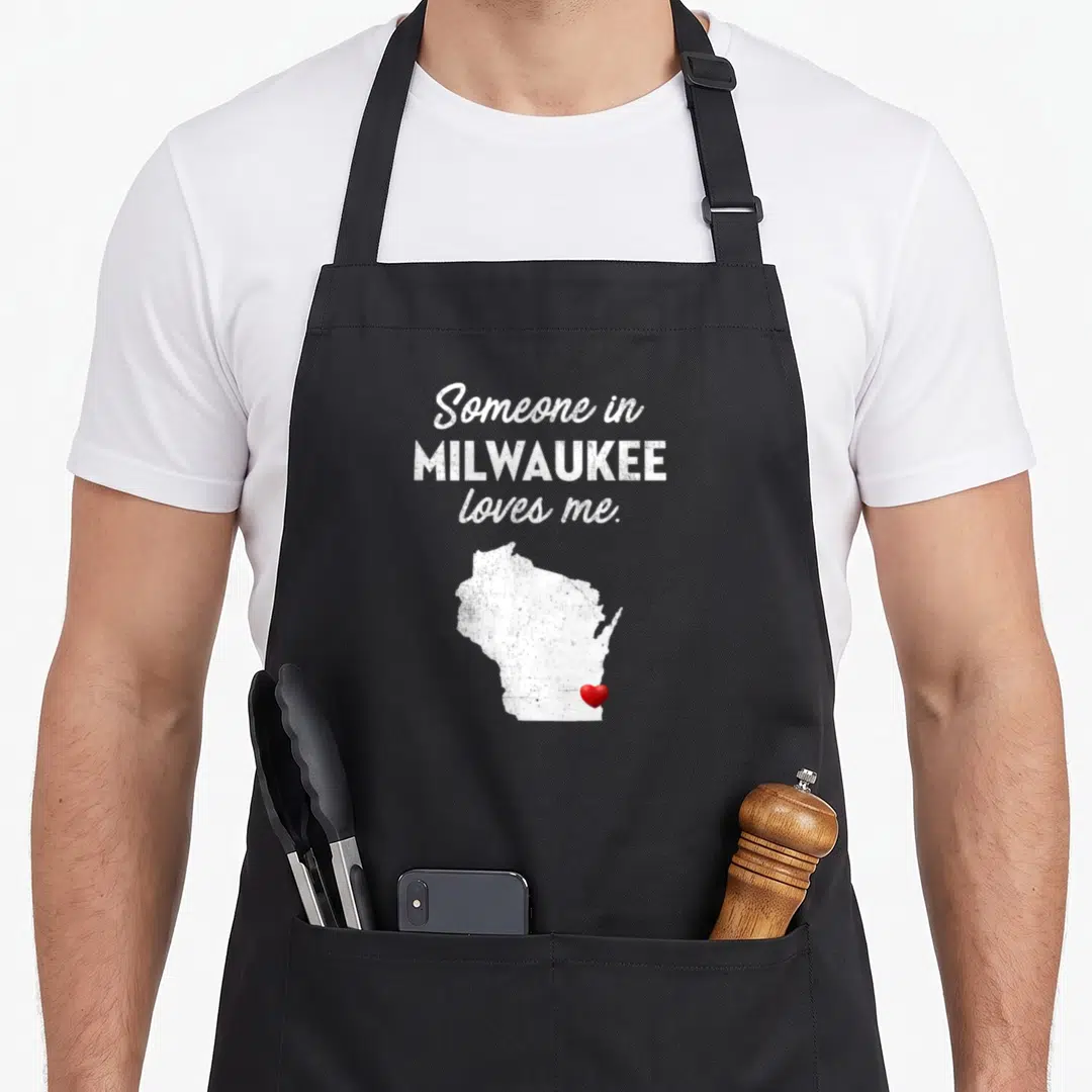Someone milwaukee apron - Distressed white Wisconsin map with red heart and text reading Someone in MILWAUKEE loves me — shown on a black cotton canvas apron against a white background