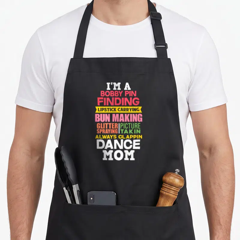Ballet mom performance apron - Colorful stacked text reading I'm a bobby pin finding lipstick carrying bun making — shown on a black cotton canvas apron against a white background