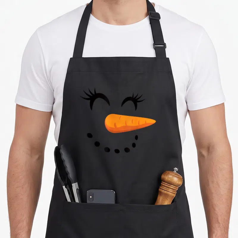Eyelash snowman apron - Cartoon snowman face with black eyelashes, orange carrot nose, and dotted smile on black — shown on a black cotton canvas apron against a white background