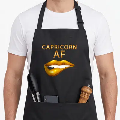 Capricorn lip birthday apron - Gold CAPRICORN AF text above cartoon lips showing white teeth — shown on a black cotton canvas apron against a white background