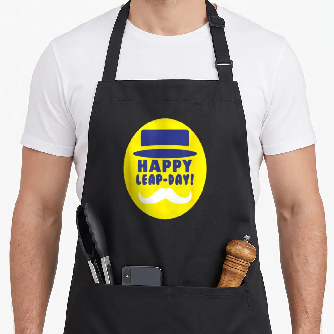 Funny leap day apron - Yellow oval with blue top hat, white mustache, and blue HAPPY LEAP-DAY text — shown on a black cotton canvas apron against a white background