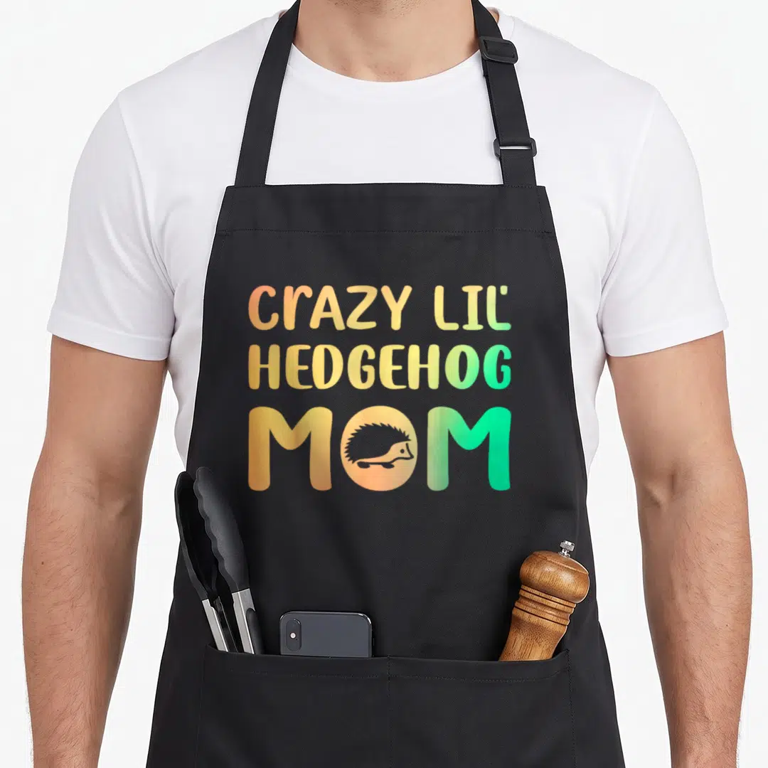 Funny lil crazy hedgehog lady apron - CRAZY LIL HEDGEHOG MOM text with hedgehog silhouette inside the O in orange — shown on a black cotton canvas apron against a white background