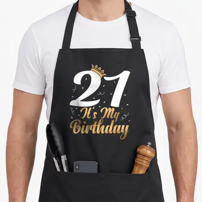 Ist funny mein apron - White 21 with gold crown and It's My Birthday script with confetti — shown on a black cotton canvas apron against a white background