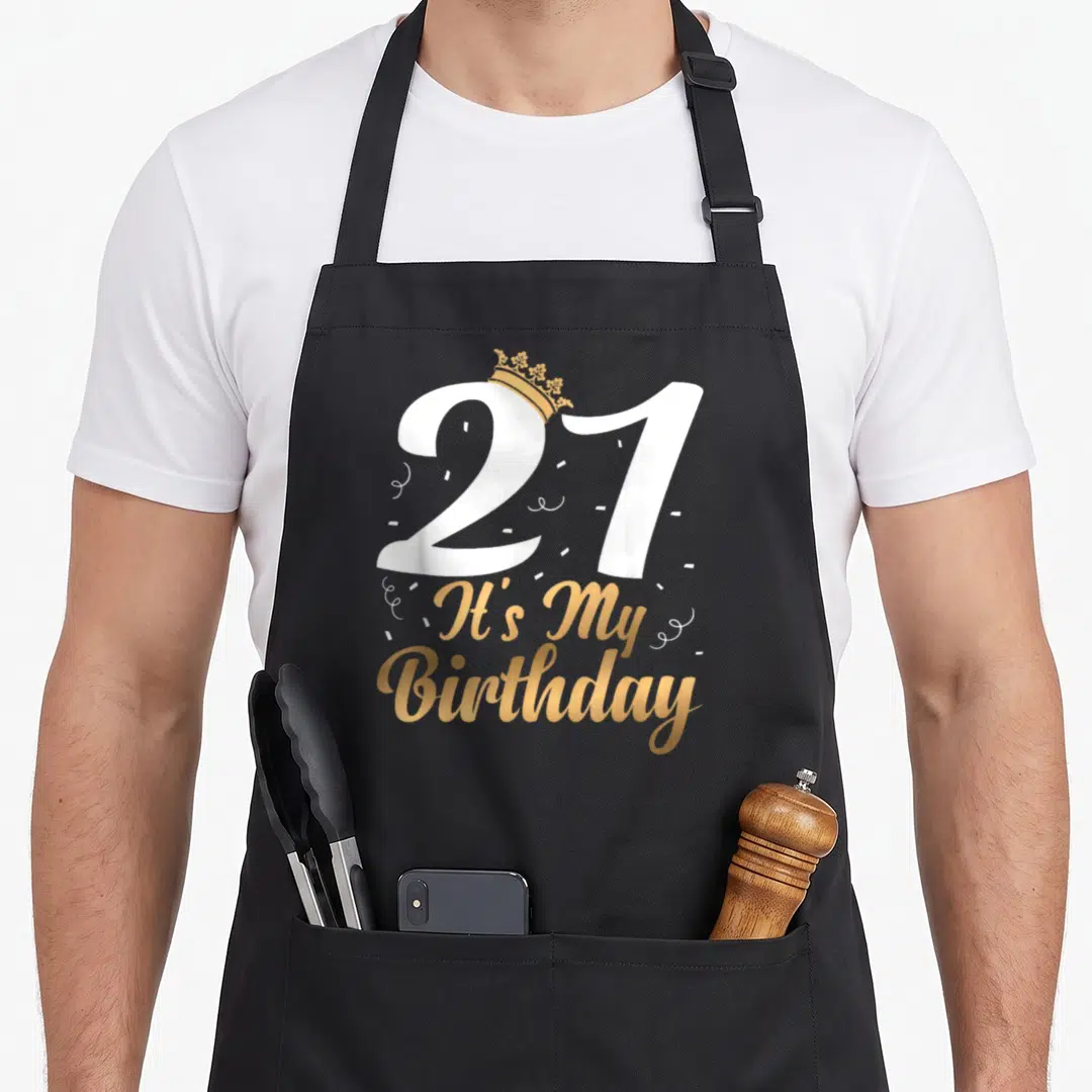 Ist funny mein apron - White 21 with gold crown and It's My Birthday script with confetti — shown on a black cotton canvas apron against a white background